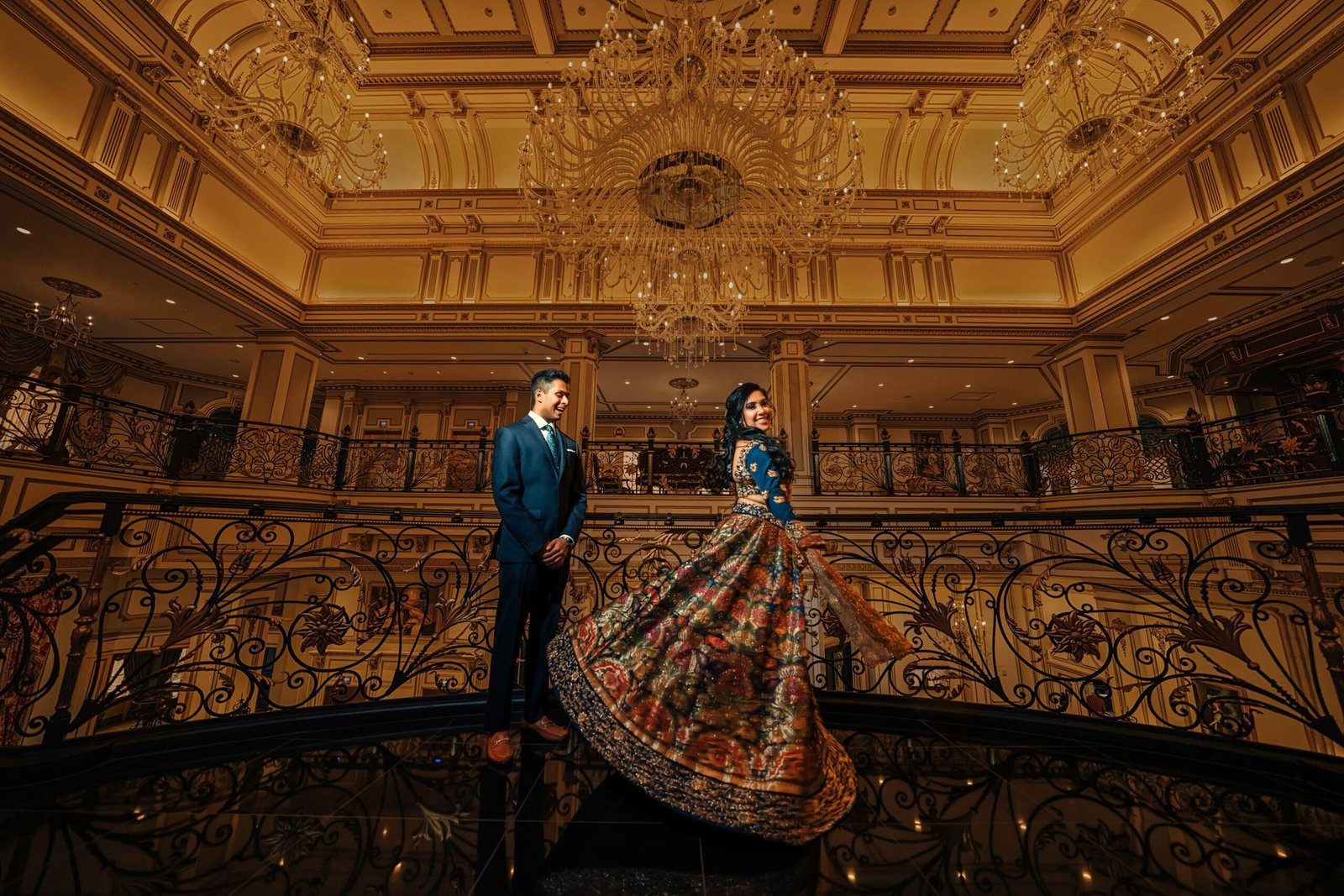 Couple in a grand gold ballroom, the bride's lehenga fanned out on the staircase